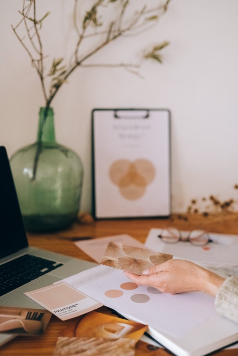 Dry plant decor in see-through green tinted glass jug next to clipboard with 3 overlapping circles of colours sitting upright leaning against the wall on a table. The table is filled with papers that have color schemes an branding exercises and a laptop.