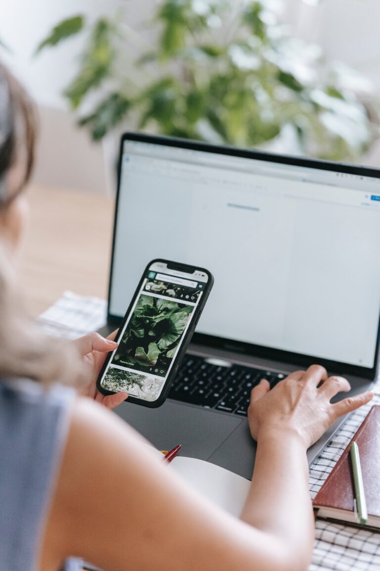 woman working on her laptop while also looking at her cellphone with a green house plant in the background